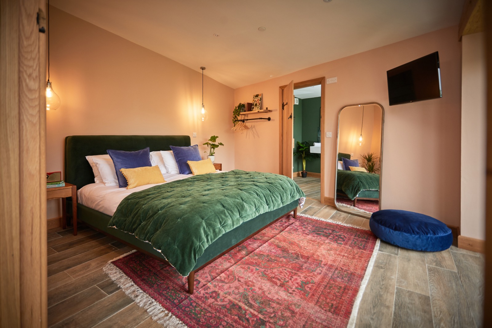 Cozy bedroom perfect for autumn getaways, featuring a green bed, red patterned rug, wall-mounted TV, and a large mirror reflecting the room’s inviting warmth.