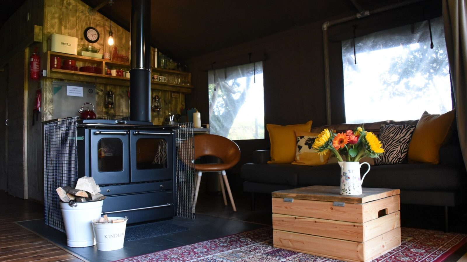 Cozy room at Stellar Safari Lodge with a stove, wooden crate table, sofa, rug, and flowers in a jug. Warm lighting and minimalistic decor create the perfect lodge ambiance.