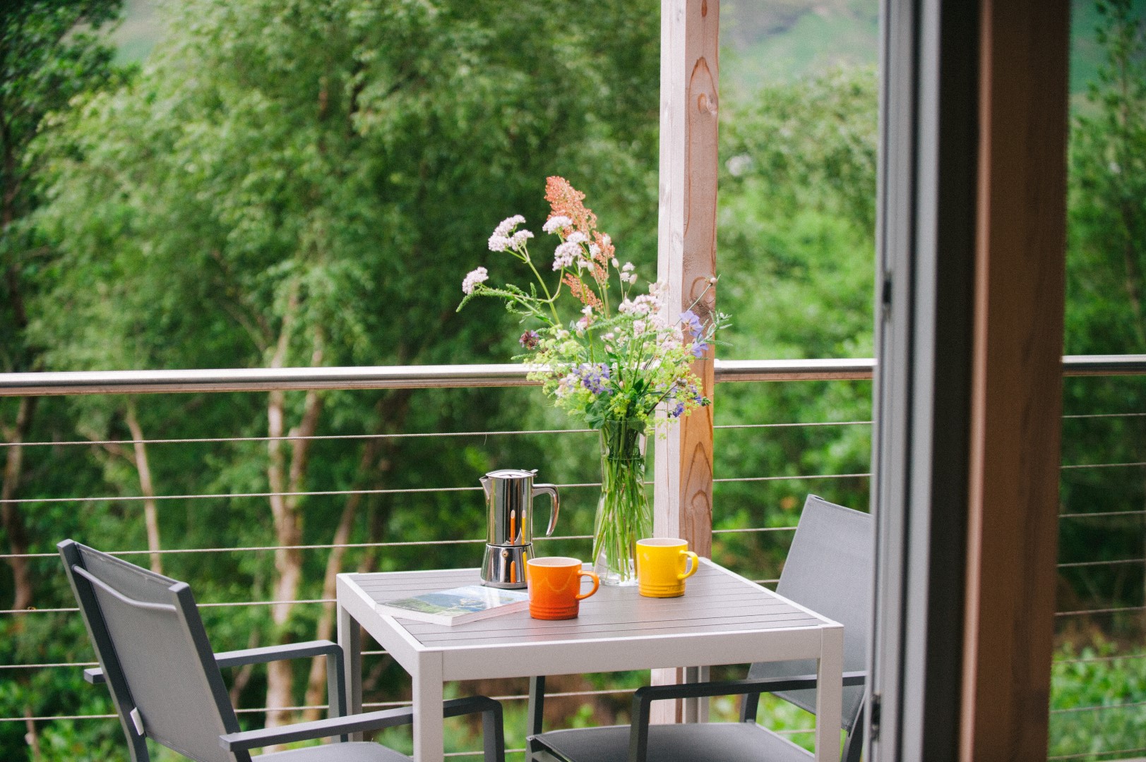 Patio table with two chairs, a coffee pot, two mugs, a magazine, and a vase with flowers. Lush greenery in the background evokes the tranquility of a greener, eco-friendly UK holiday.