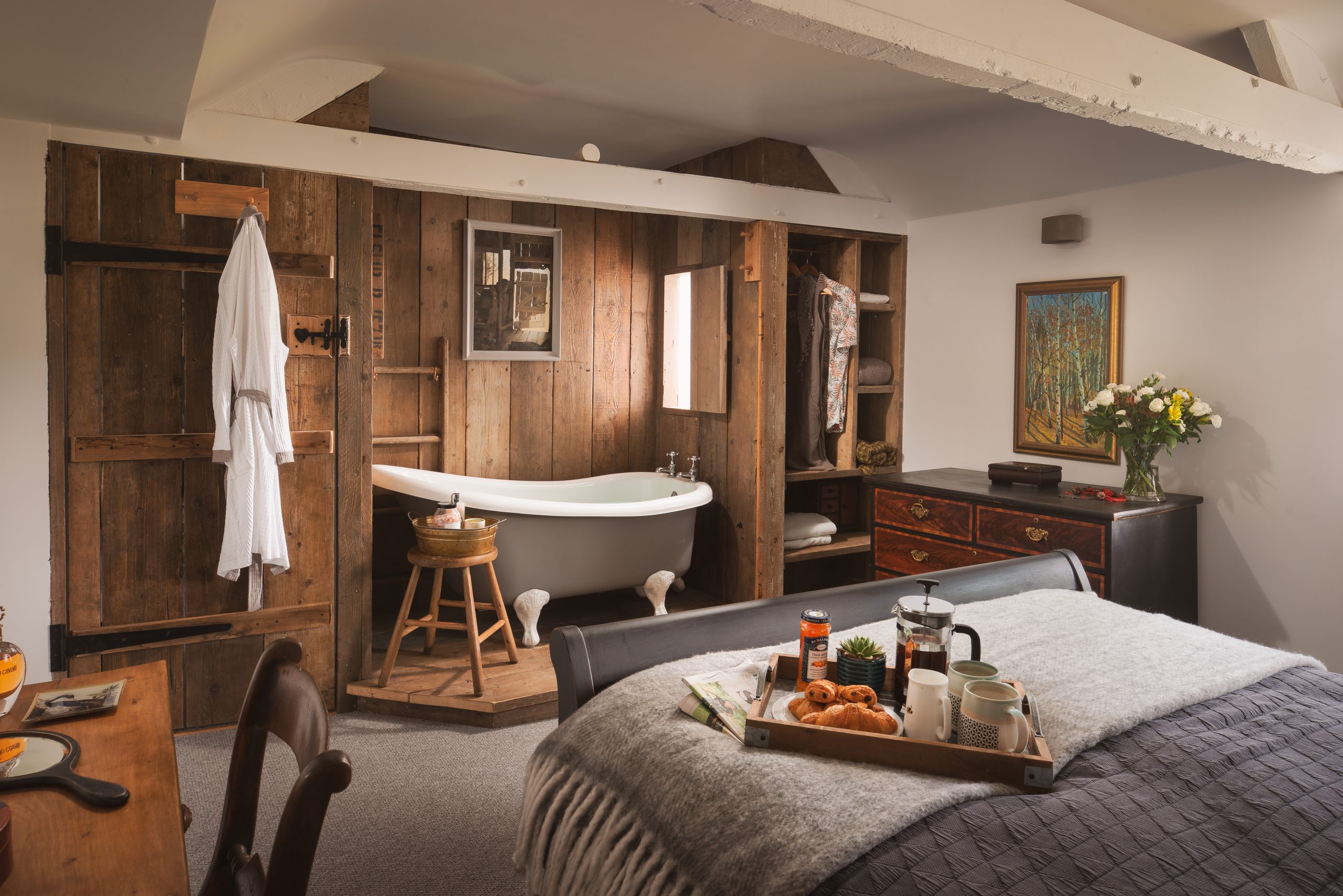 Nestled within the charm of rustic wooden walls, this cozy bedroom is your serene escape in one of 8 Cosy Cottages. Unwind with a relaxing soak in the vintage bathtub, then snuggle up under a gray blanket, indulging in breakfast delights conveniently placed on a tray nearby.