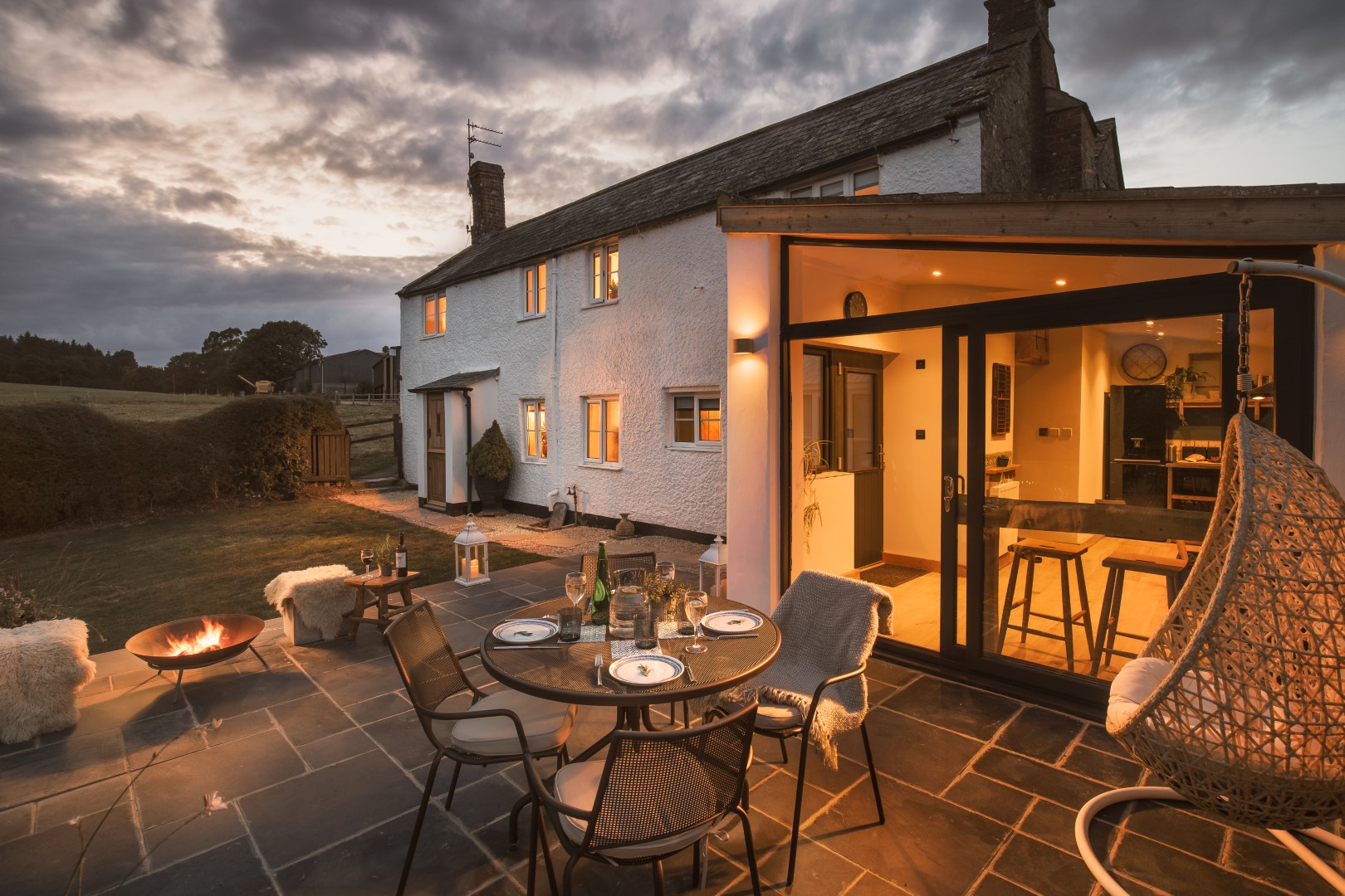 A cosy outdoor patio features a dining table set for four, a fire pit, and a swinging chair. Nestled beside one of 8 Cosy Cottages, the scene is beautifully lit at dusk, exuding rustic charm.