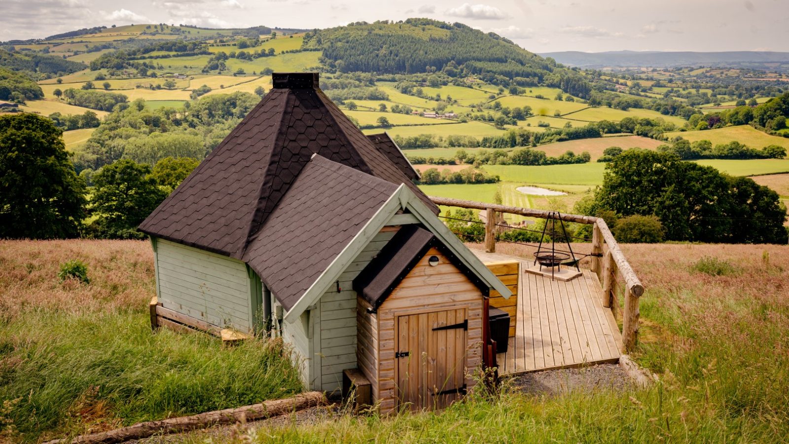 A small, rustic cabin with a dark roof sits on a grassy hillside. From the wooden deck, where chairs offer rest, one can overlook a lush green valley and rolling hills that seem drawn from the pages of a fantasy novel under a partly cloudy sky.