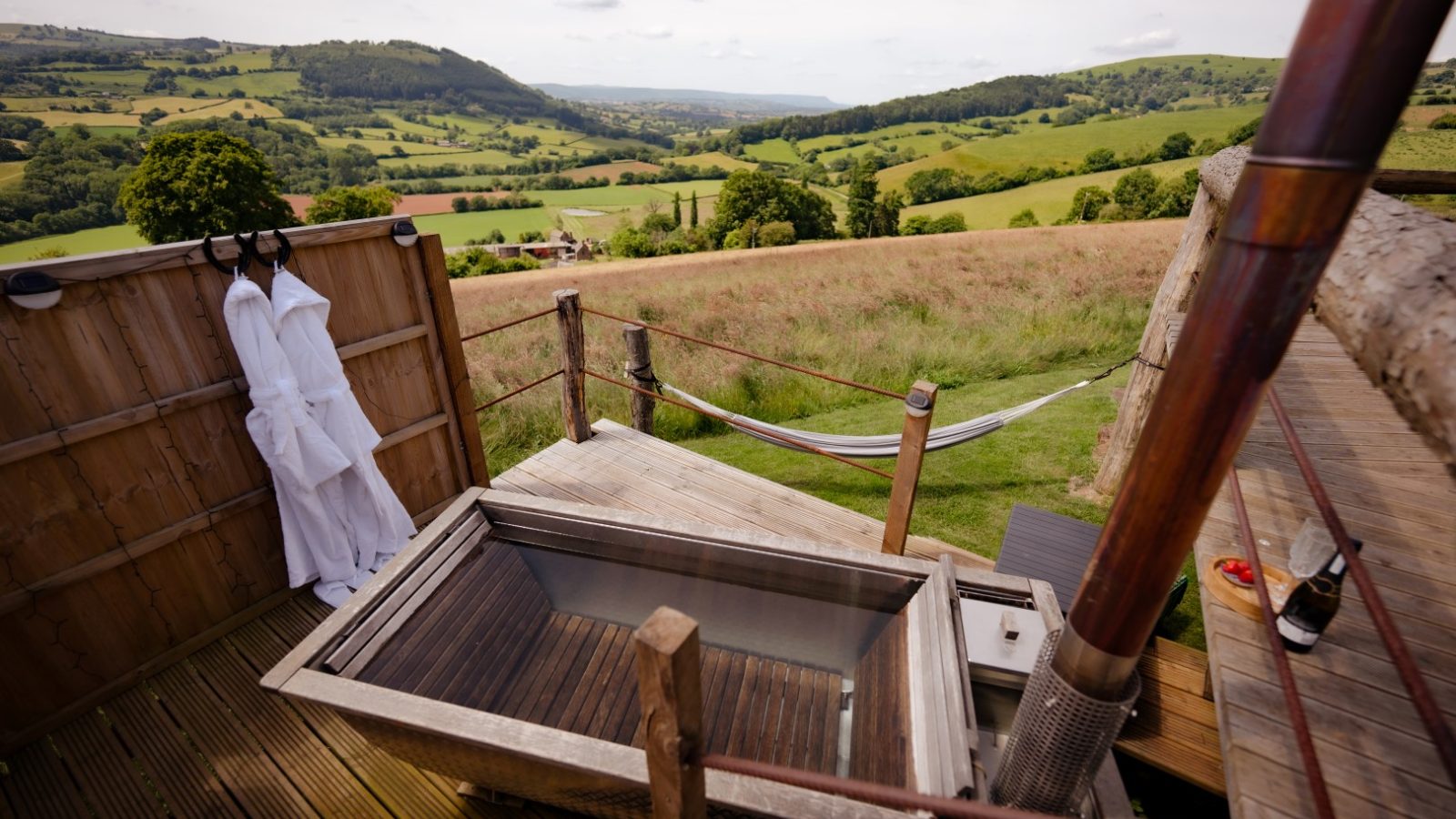 A wooden outdoor bath on a deck overlooks the lush green valley of Dragons Gate, with rolling hills and scattered trees. Two white robes hang on the wall, and a striped hammock is set up nearby. A partially cloudy sky adds to the serene atmosphere.