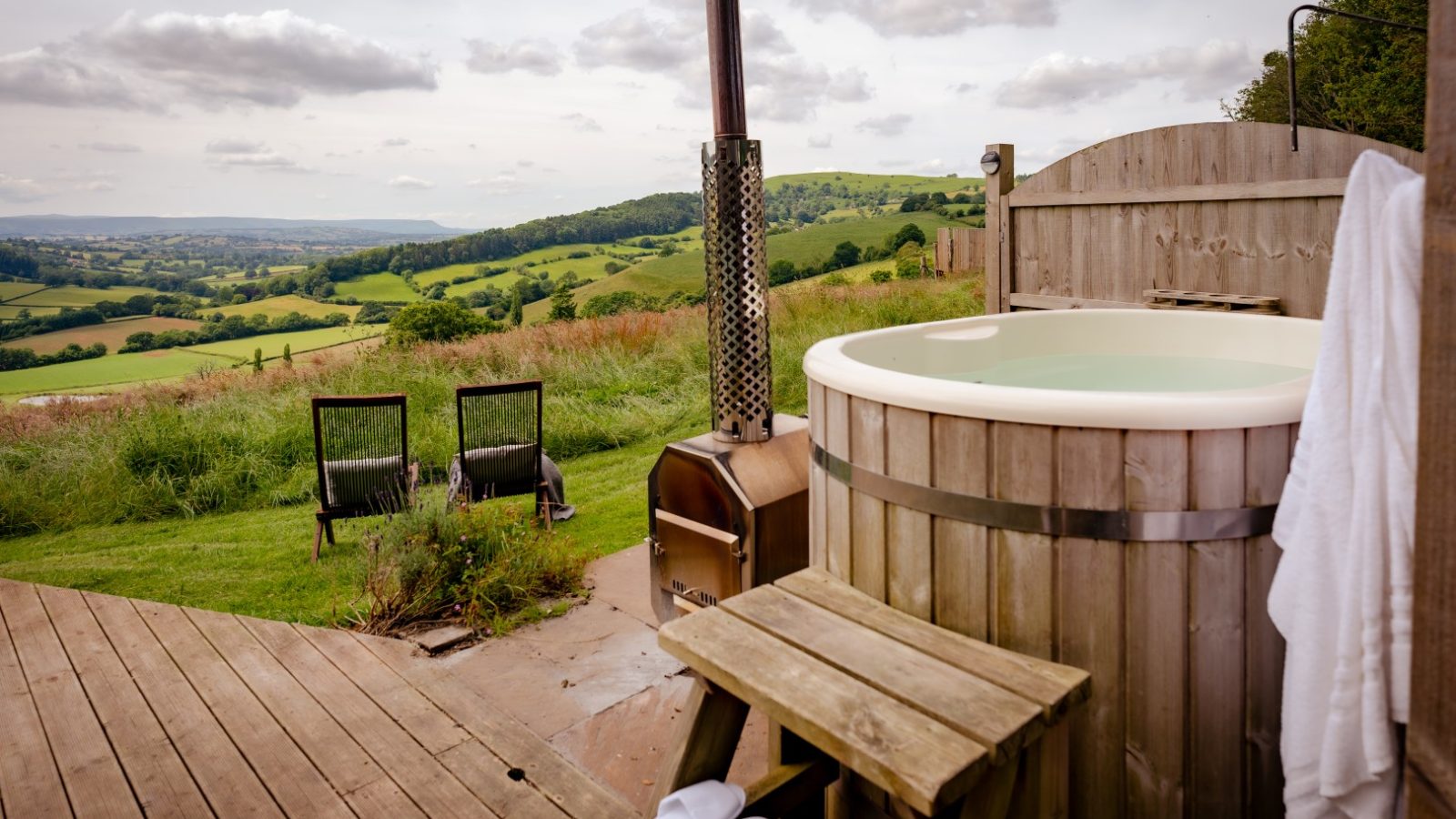 A wooden hot tub rests on a patio with two chairs, offering a tranquil view of the scenic countryside. Under a partly cloudy sky, the enchanting backdrop evokes a sense of mystery as if guarding the serene beauty like an ancient Dragons Gate.