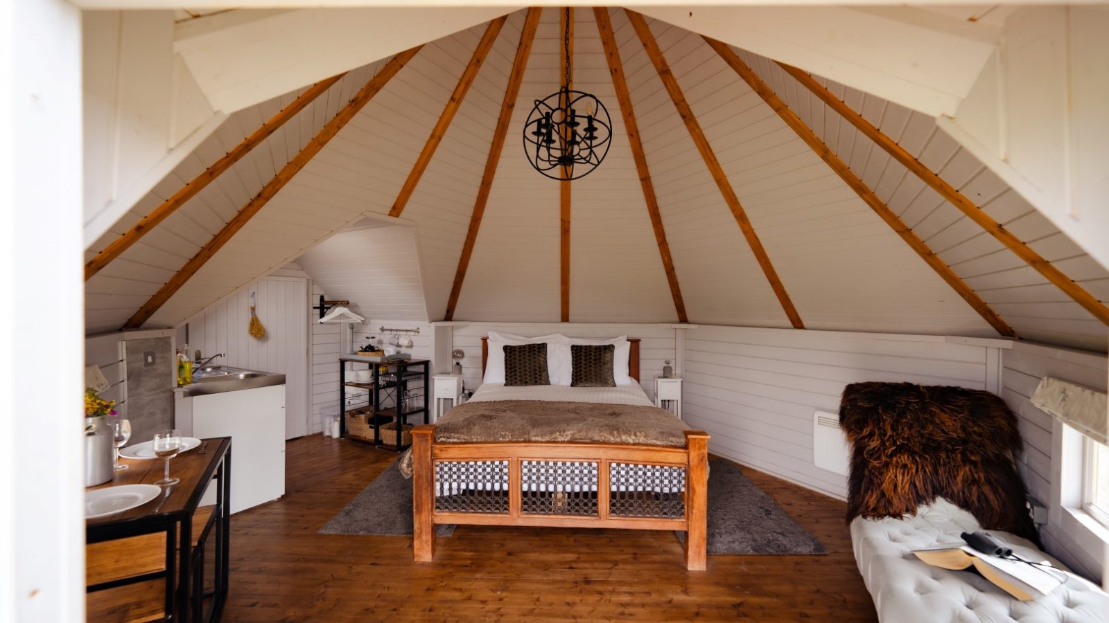 Cosy triangular cabin interior with a bed, kitchenette, and seating area. The ceiling boasts a rustic wood beam pattern. Dragons Gate Glamping Cabins in Herefordshire