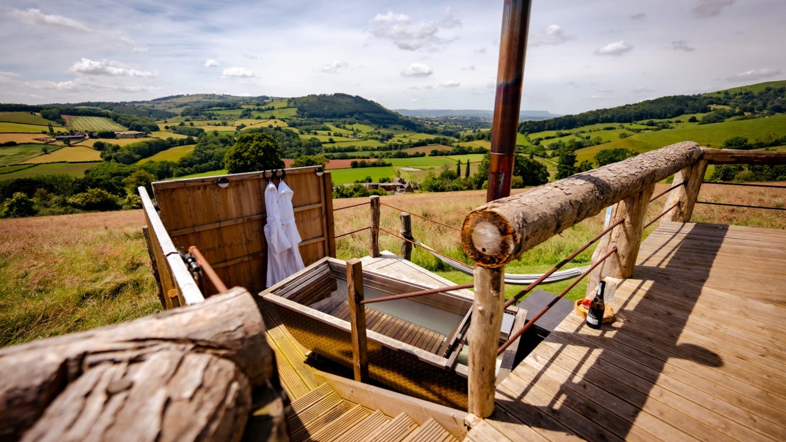 An outdoor bathing area with a standalone tub on a wooden deck overlooks rolling green hills and a cloudy sky, reminiscent of a mystical Dragons Gate. Two white robes hang on a wooden screen for privacy. A hammock and deck chairs complete the rustic setting.