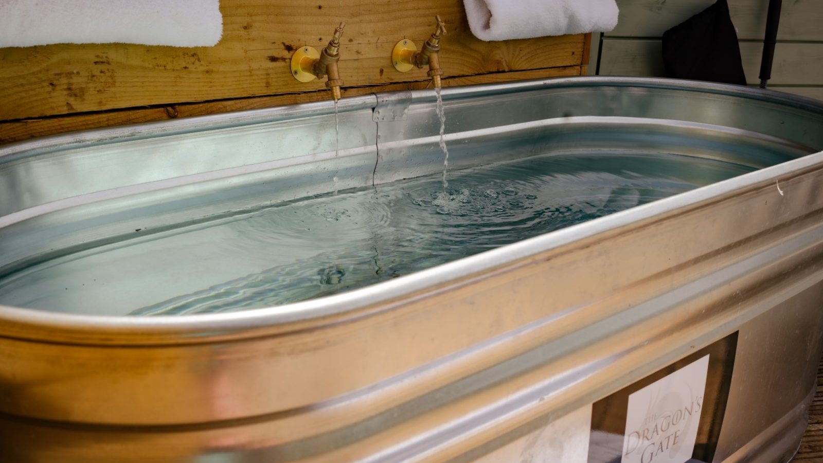 A large metal tub filled with clear water stands outdoors beneath the Dragons Gate, under two running taps. Two white towels are neatly placed on the wooden board above the tub.