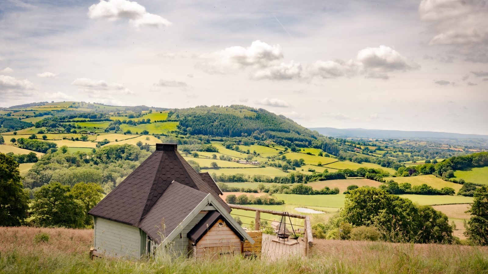 A small cabin with a sloped roof sits in the foreground, surrounded by grass. Beyond it lies Dragons Gate, where rolling green hills and farmland stretch under a partly cloudy sky, creating a serene rural landscape.