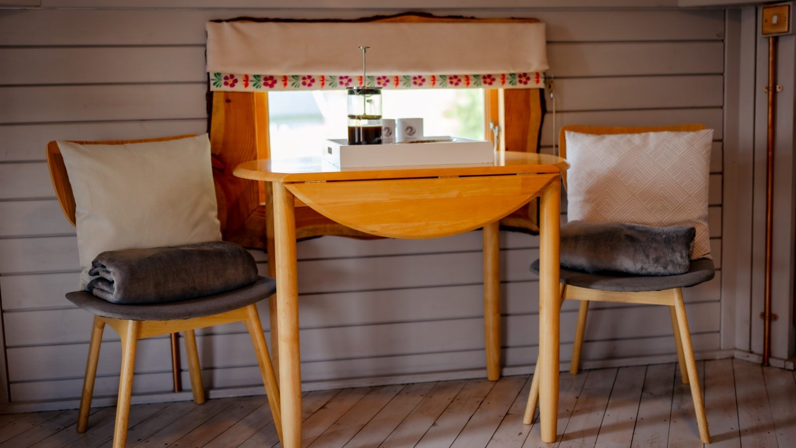 Cosy cabin corner with a wooden table, two chairs adorned with cushions, and a French press alongside mugs on a tray by the window—a perfect spot to relax or dive into a novel. Dragons Gate Glamping Herefordshire