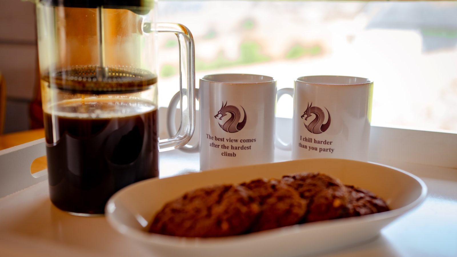 A French press coffee, two mugs with unicorn emblems, and a plate of cookies sit on a white tray by the window, offering a serene view from the cabin at Dragons Gate Glamping