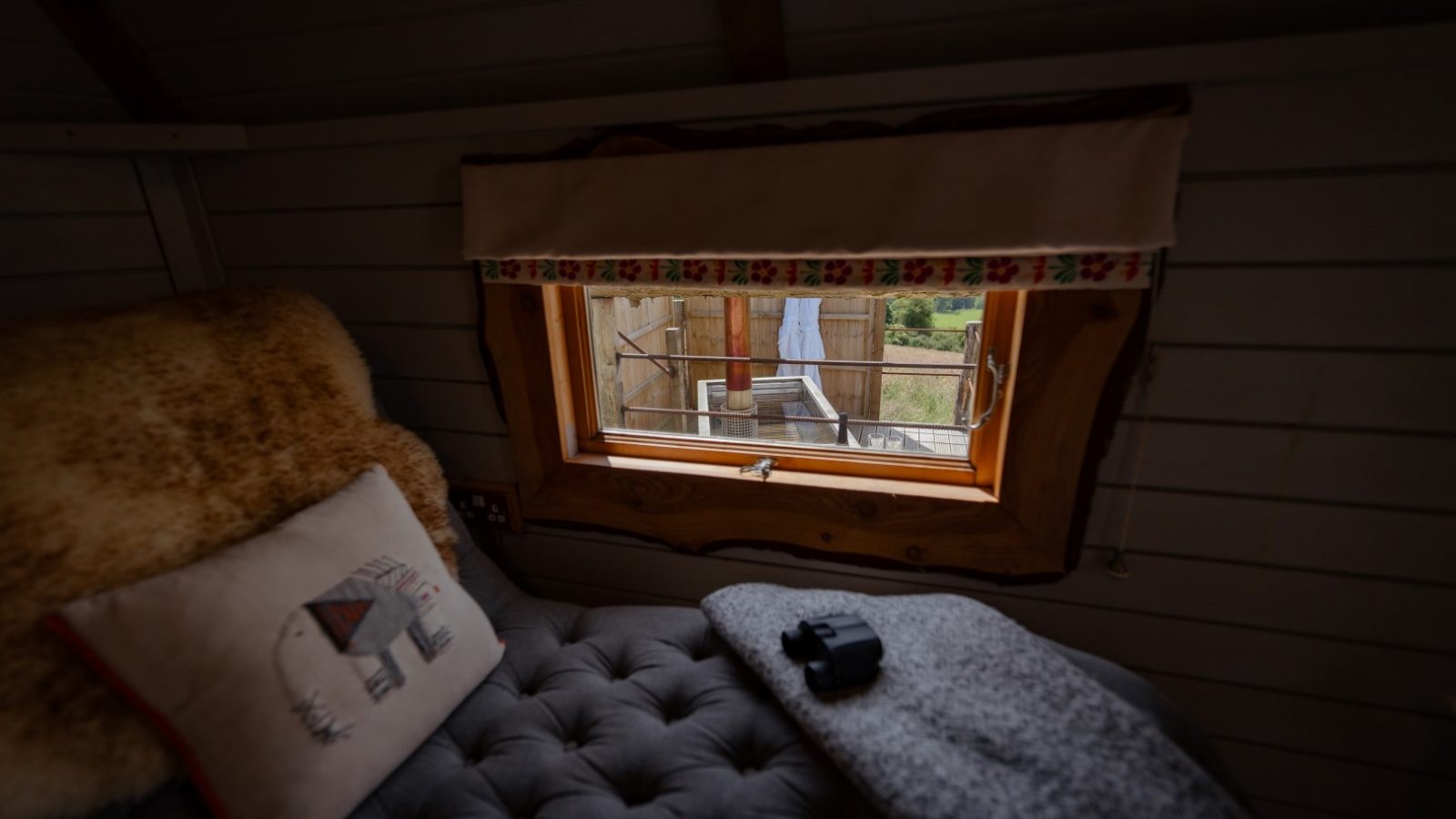 Cozy cabin interior with a tufted couch, pillow, and window view of the Dragons Gate countryside. A camera rests on a blanket nearby.