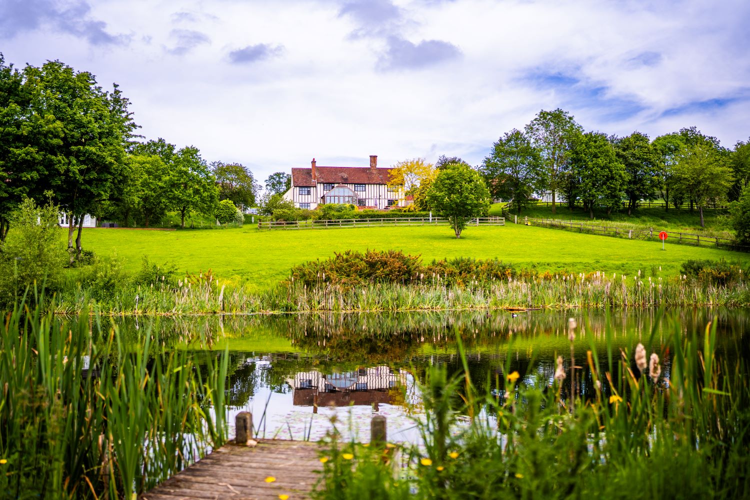 Perched on a hill, this large white house offers a serene retreat, boasting a lush green lawn, trees all around, and a tranquil pond with a dock. Nearby, 8 cosy cottages blend seamlessly into the picturesque landscape for an enchanting escape.