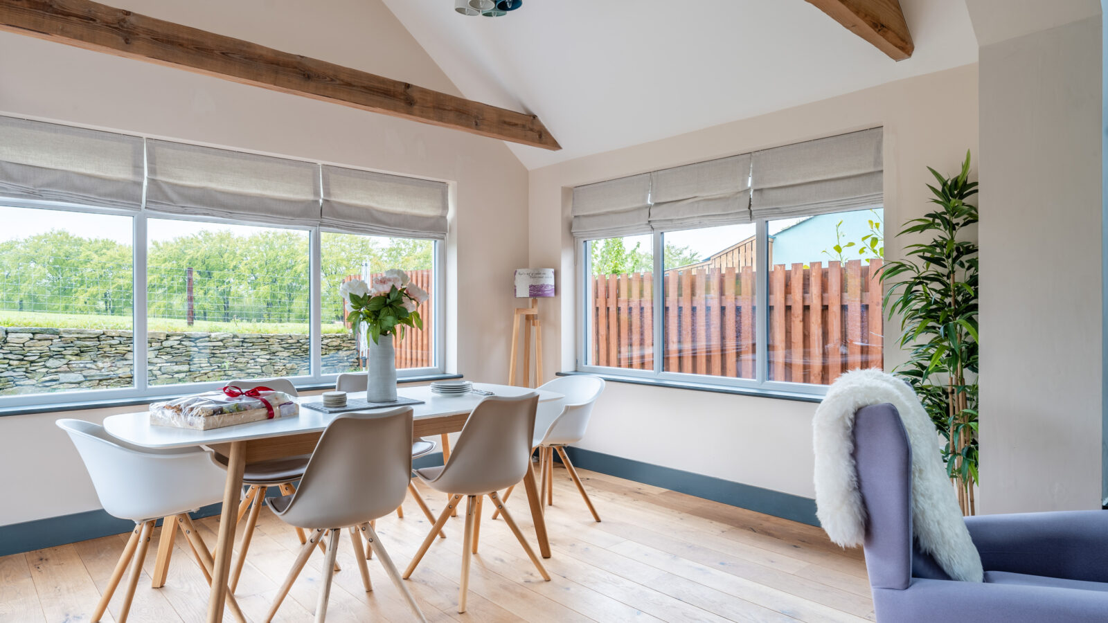 The bright Stabl Nantcoy dining room boasts wooden beams, a table with chairs, and a potted plant. Large windows overlook a garden and fence, infusing the space with tranquility.