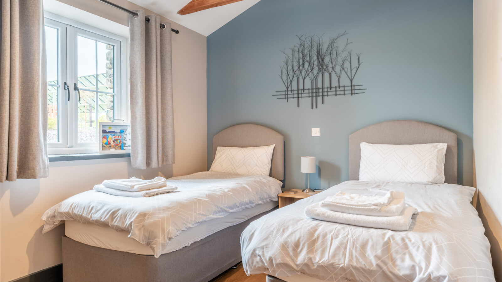 A bedroom at Stabl Nantcoy features two single beds with gray headboards, a small nightstand, a lamp, and wall art adorning a blue-gray accent wall.