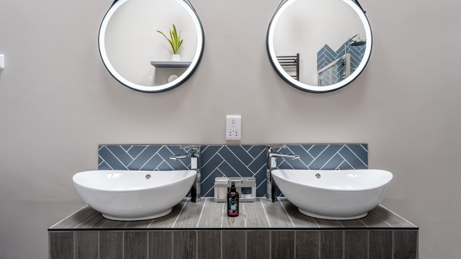 A modern bathroom featuring two round mirrors, two vessel sinks on a Stabl Nantcoy wooden countertop, and a herringbone tile backsplash.