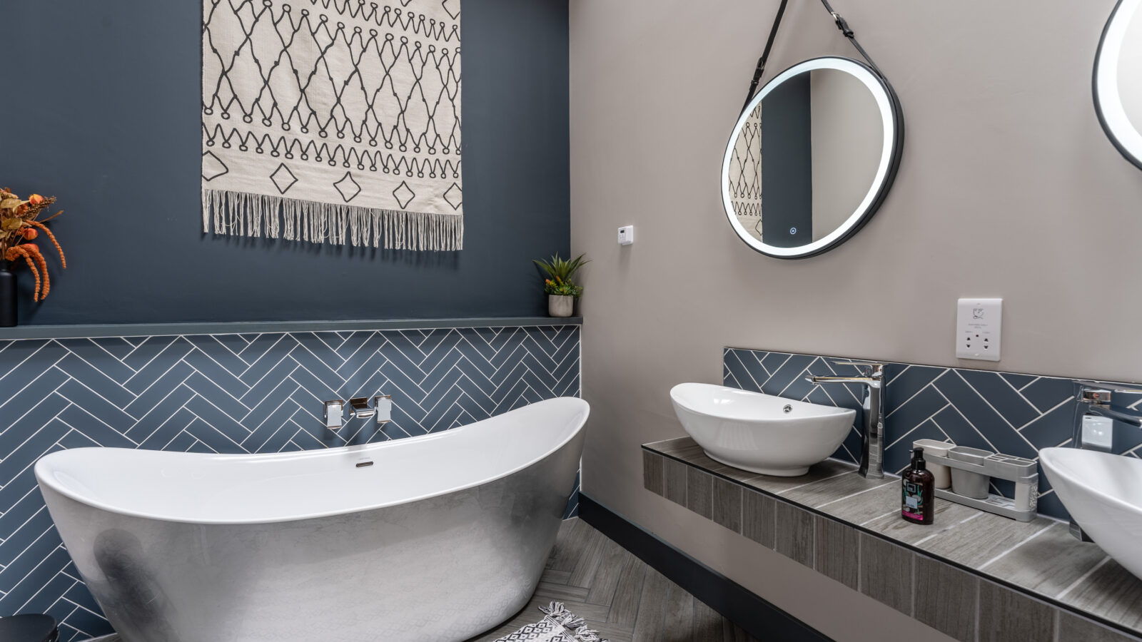 Modern bathroom with a freestanding tub, round mirrors, dual sinks, geometric wall tiles, and a decorative Stabl Nantcoy wall hanging.