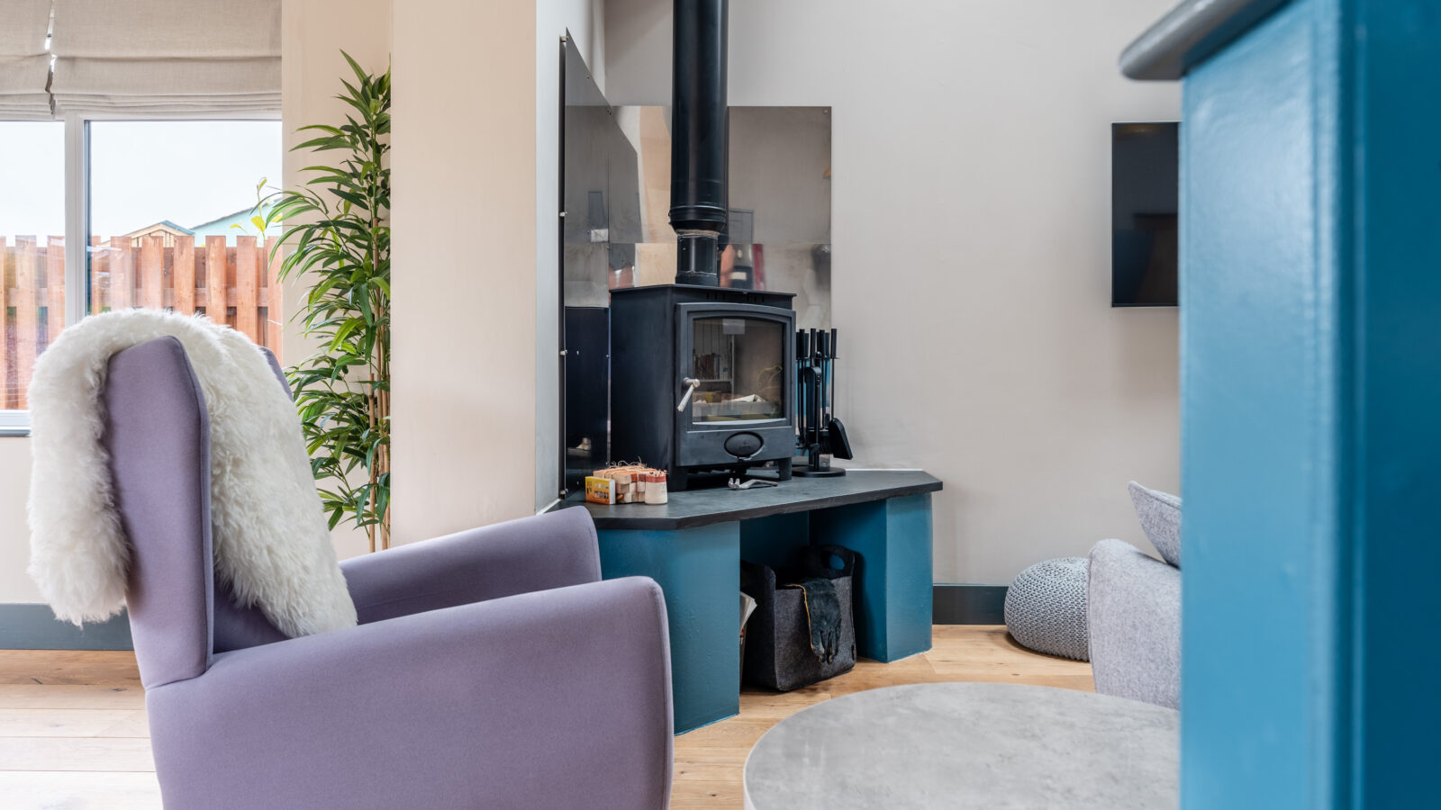 A cozy living room at Stabl Nantcoy features a purple armchair, a wood-burning stove on a blue hearth, and a small table in the foreground.