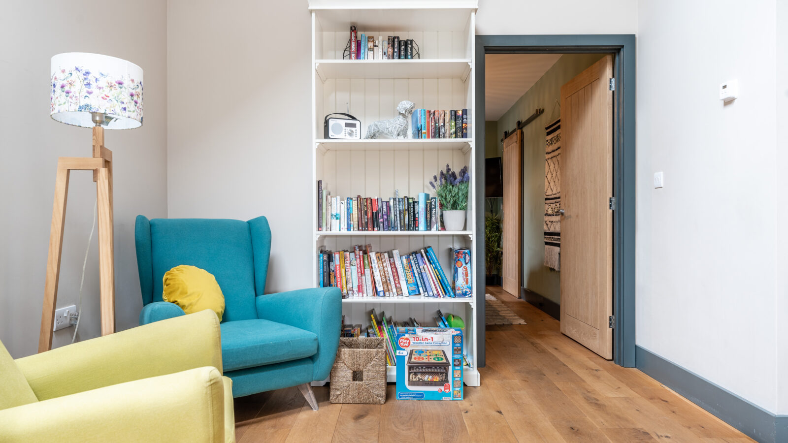 The living room features a white bookshelf, teal and yellow armchairs, a floral lamp, and a polished wooden floor. A door gracefully opens to the hallway of Stabl Nantcoy.