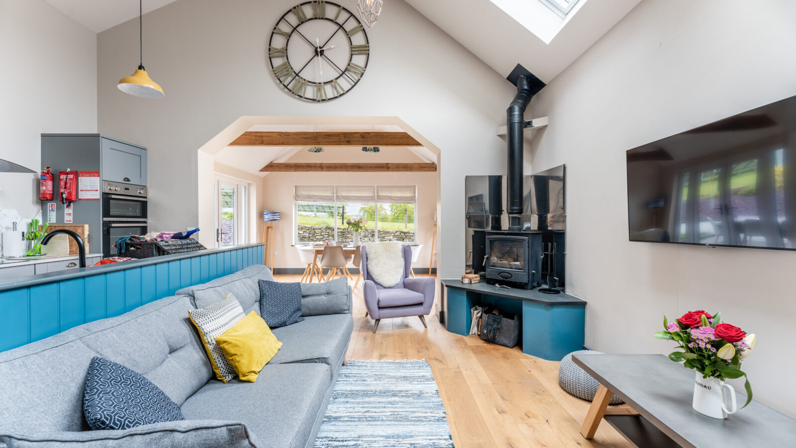 A modern living room in Stabl Nantcoy features a large gray sofa, wall clock, wood stove, skylight, and wall-mounted TV. Flowers elegantly adorn the coffee table.