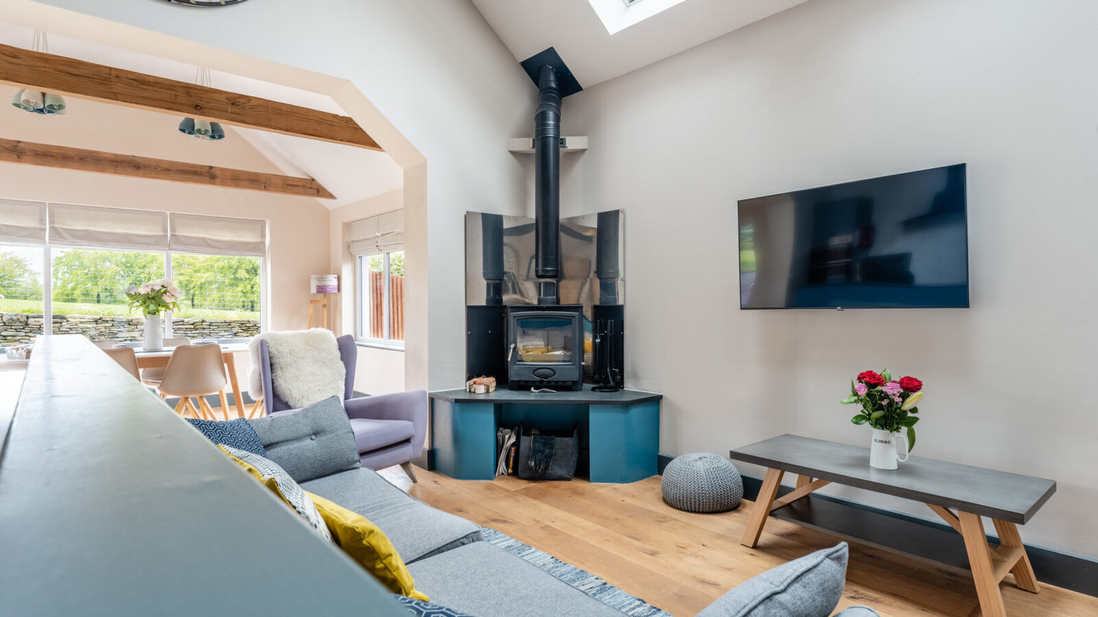 Bright living room with a wood-burning stove and a wall-mounted TV, accented by the rustic charm of Stabl Nantcoy's wooden beams. A vase with roses graces the coffee table. 