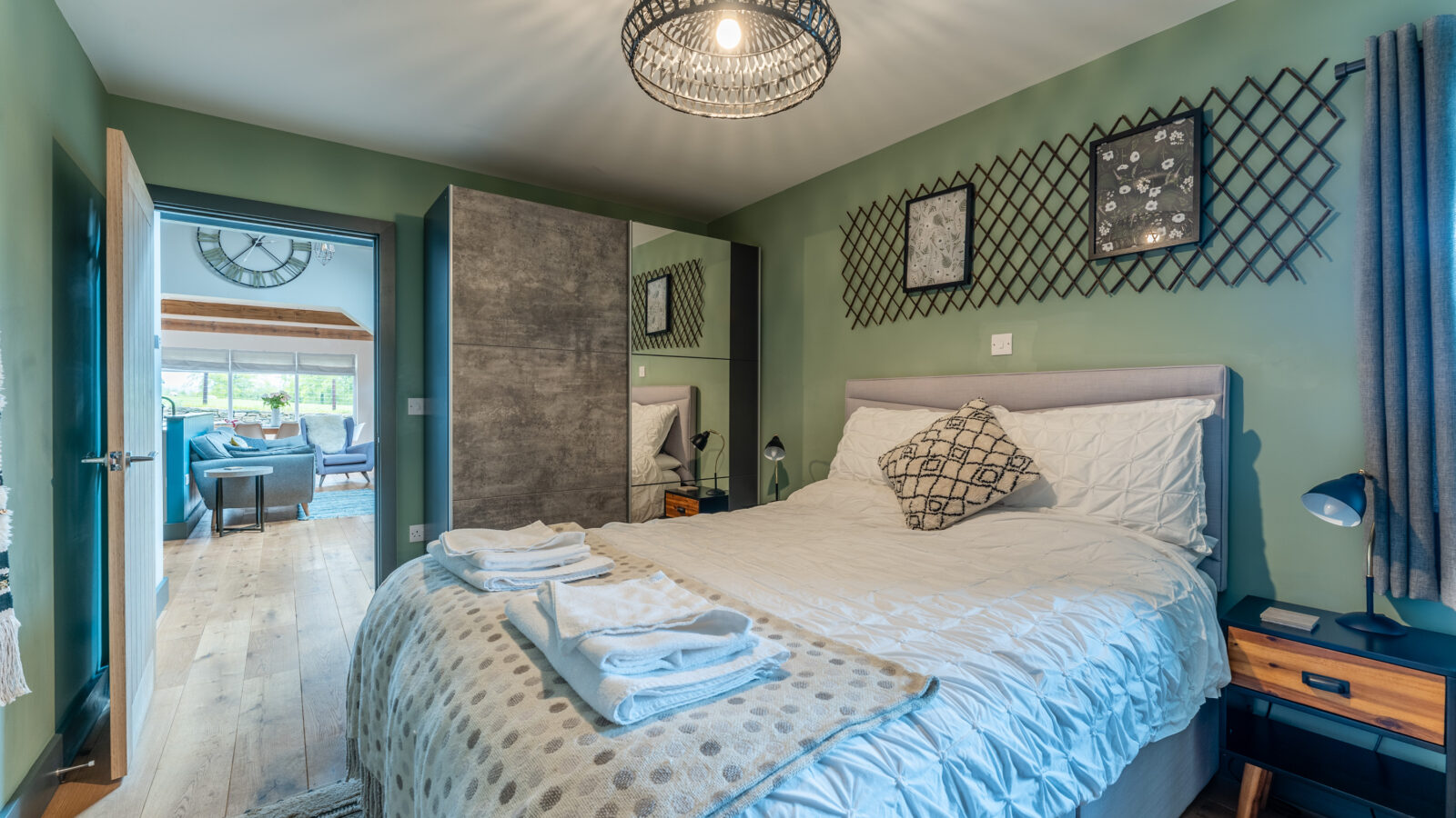 A bedroom at Stabl Nantcoy features a bed, modern decor, cozy folded towels, and an open door leading to a living area adorned with a striking large clock on the wall.