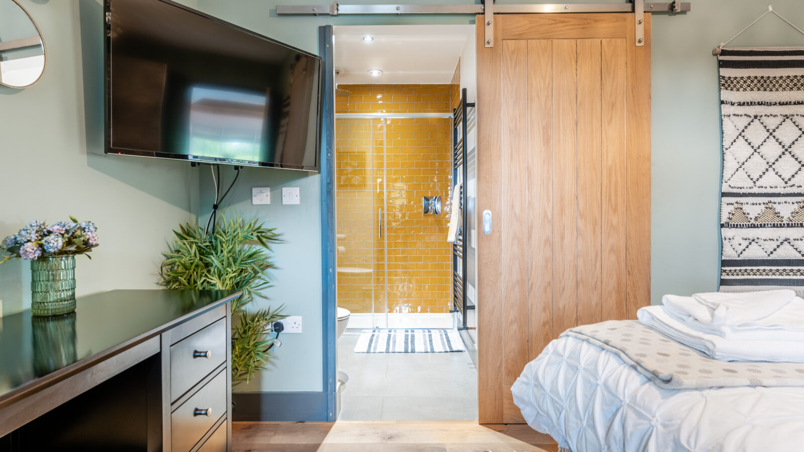 The bedroom features a wall-mounted TV and a sliding door that reveals a bathroom with charming Stabl Nantcoy yellow tiles. The bed is neatly made, inviting comfort and relaxation.