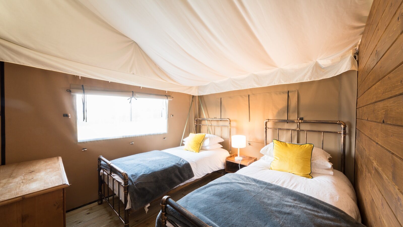 Interior of a Nest Glamping tent with two single beds, metal frames, yellow pillows, and a small wooden nightstand with a lamp.