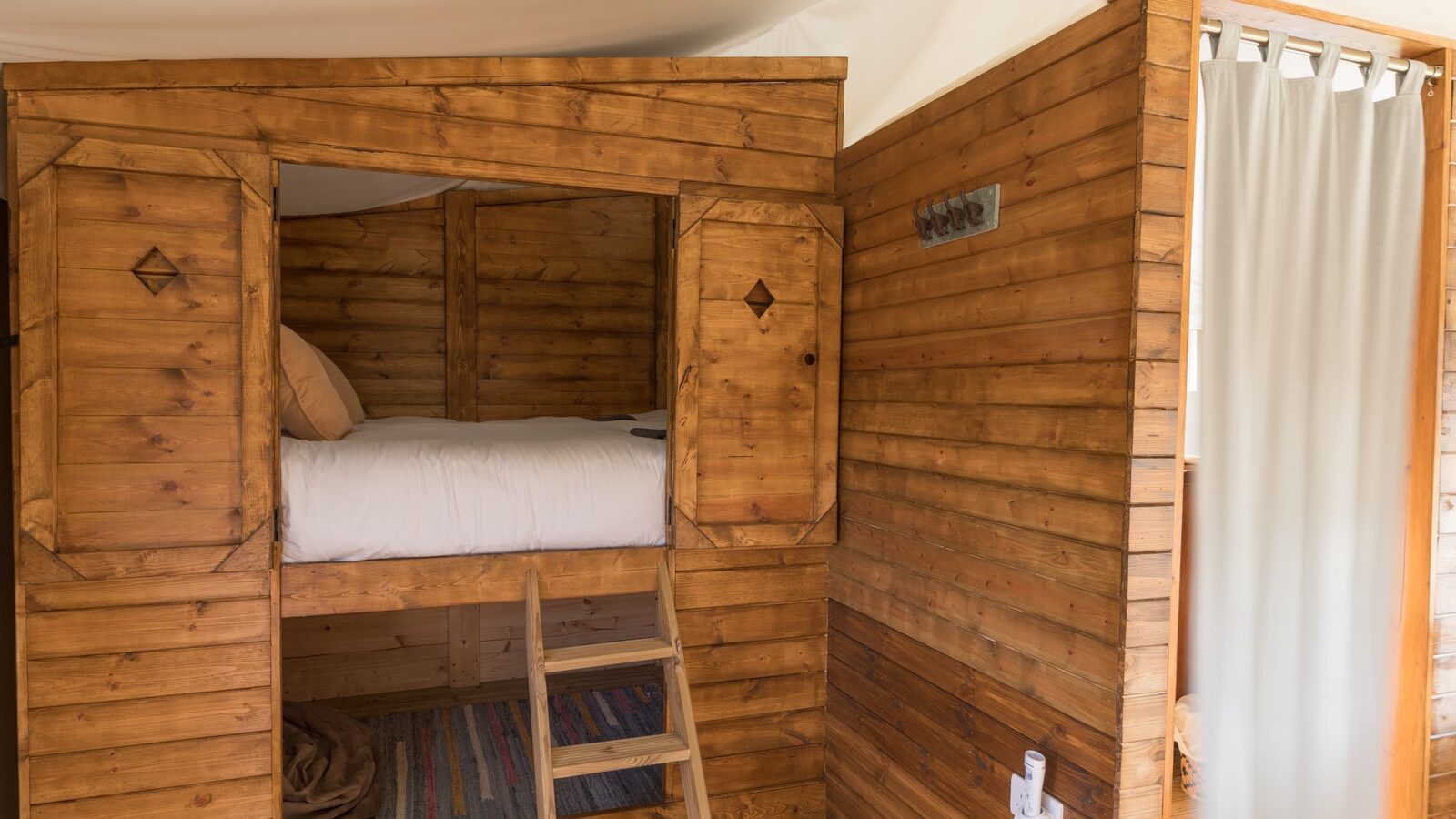 Nestled within a cozy wooden interior, this luxury camping spot features a bunk bed accessible by ladder and a small adjacent bathroom area discreetly concealed by a curtain for privacy.