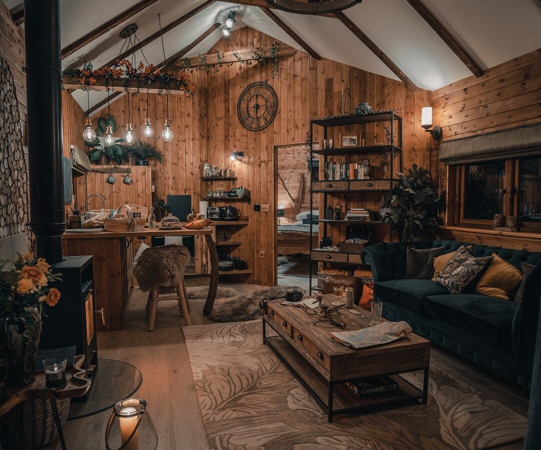 Cozy wooden cabin interior reminiscent of a Woodland Chase, featuring a couch, kitchen, table, and hanging chandelier. Decor includes clock and autumn accents, creating a warm treehouse ambiance.