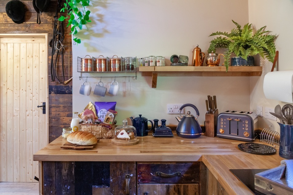 Rustic kitchen with wooden countertops, a loaf of bread, tea kettle, toaster, jars, and plants on shelves. A wooden door completes the charming space, reminiscent of a cozy treehouse nestled in Woodland Chase.