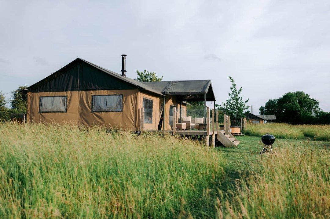 A large canvas tent with a porch elegantly nestles in a grassy field, embodying the essence of Nest Glamping. A small barbecue grill stands nearby under the cloudy sky, offering a taste of luxury camping.