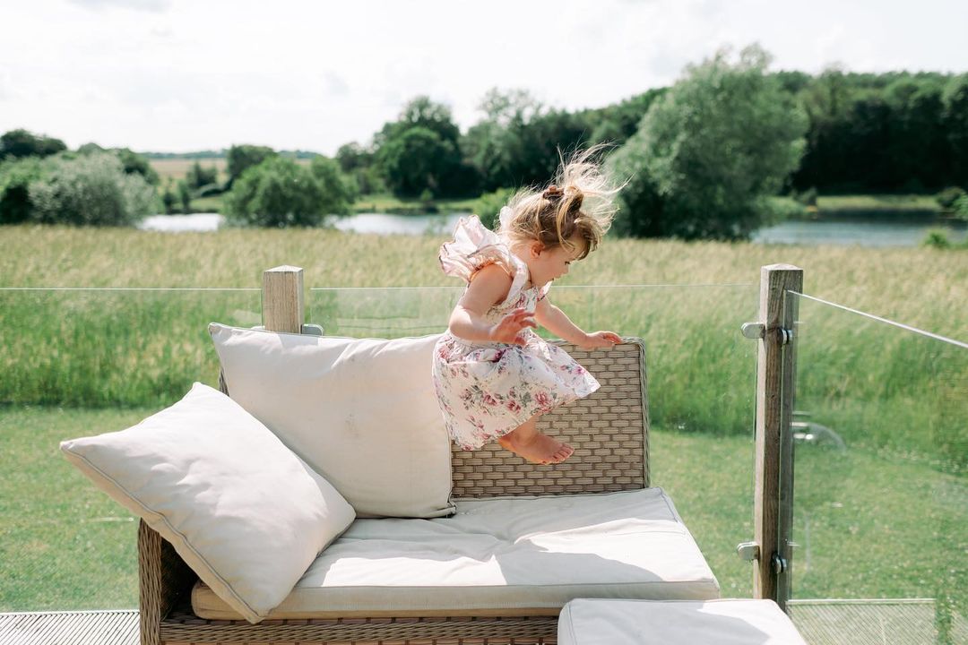 A young girl in a floral dress jumps onto a cushioned sofa on an outdoor patio, nestled amidst a grassy field and trees, capturing the essence of glamping.