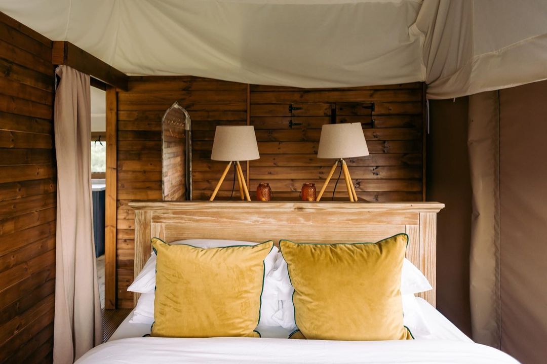 Cozy bedroom with wooden walls reminiscent of an outdoor glamping retreat, featuring a double bed, yellow pillows, and two lamps on the headboard shelf.