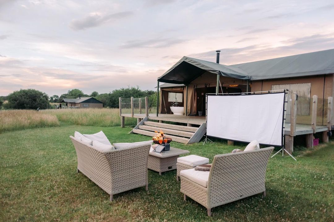 Nest Glamping offers a luxurious outdoor seating area with wicker furniture facing a blank projector screen, in front of a tented structure on a grassy field. Perfect for an unforgettable glamping experience.