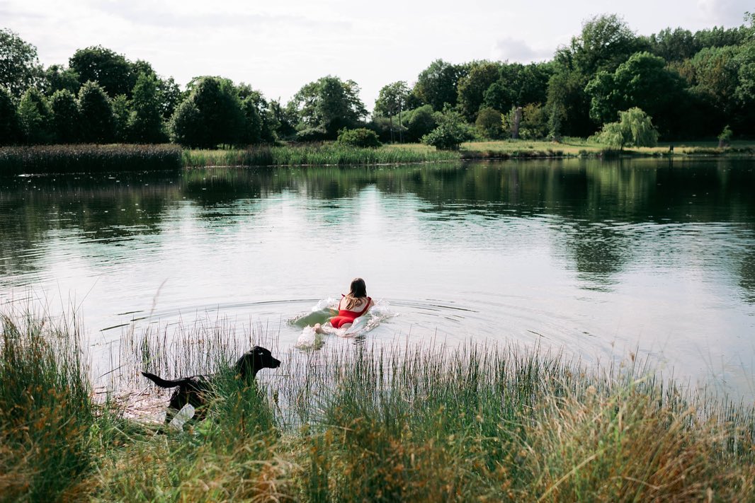 A person in a red swimsuit floats on a raft in a lake, enjoying their glamping experience, while a black dog plays near the grassy shore under the cloudy sky.