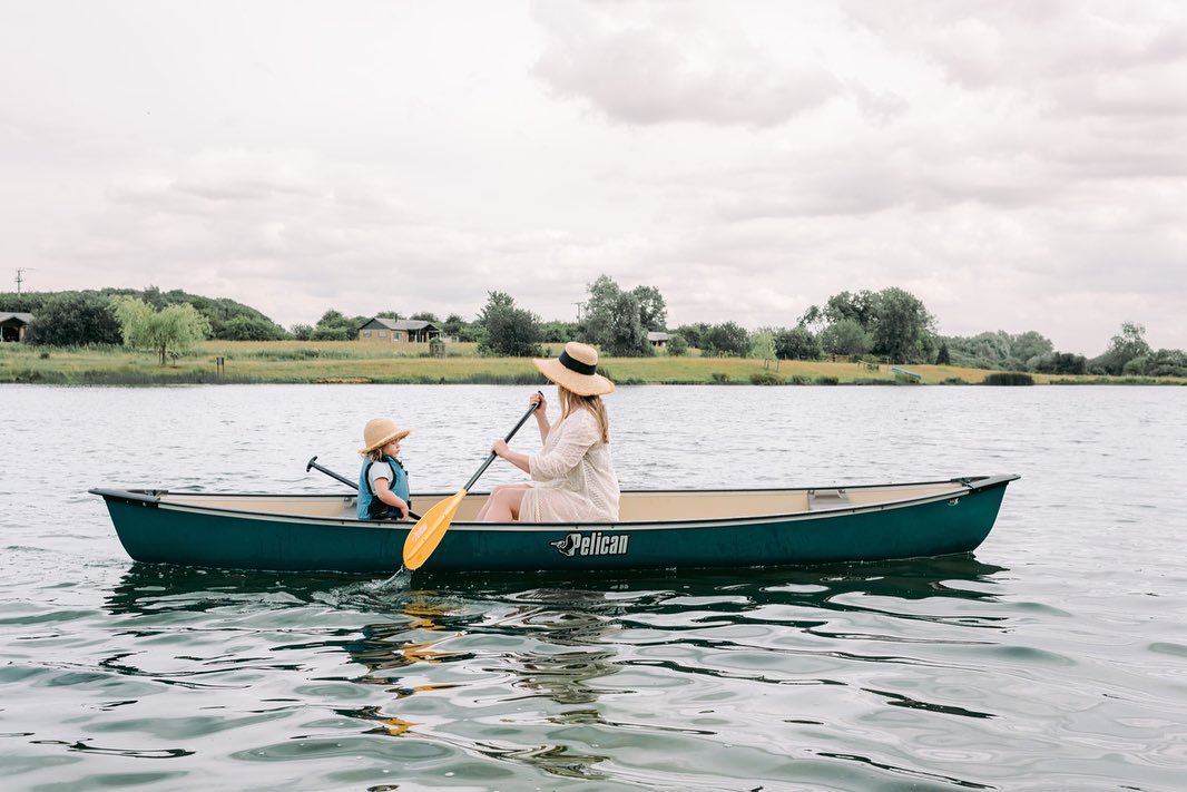 An adult and child, both wearing hats, paddle a green canoe on a calm lake with trees and houses in the background, enjoying nature's tranquility during their luxury glamping getaway.