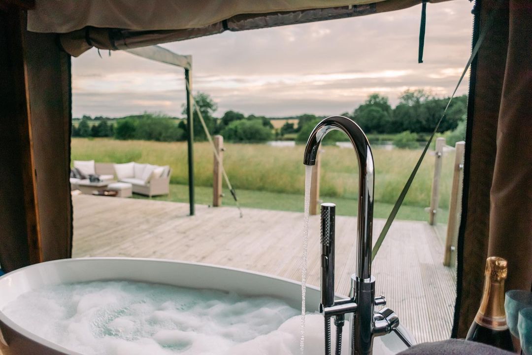 The bathtub with running water is perfect for a glamping experience, overlooking a wooden deck and green field. Enjoy outdoor furniture under the cloudy sky, epitomizing the essence of Nest Glamping.