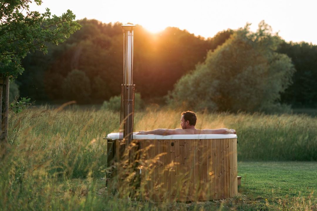 Nestled in a round wooden hot tub, enjoying the luxury of Nest Glamping, a person unwinds amidst nature's tranquility. Surrounded by lush grass and trees, they soak in the serene ambiance as the sun sets beautifully in the background.