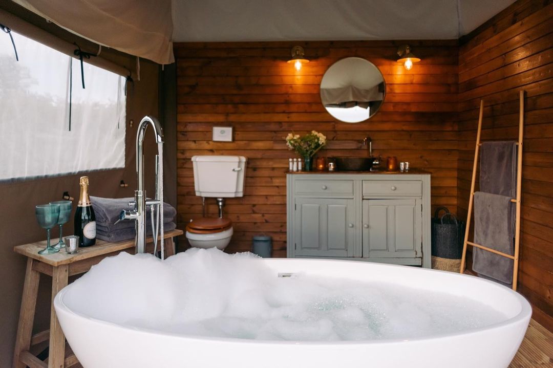 Indulge in a nest glamping experience with this wooden bathroom, featuring a bubble-filled bathtub, mirror, cabinet, and toilet. Savor the luxury with a bottle of champagne and glasses nearby.