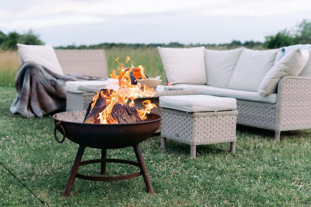 Experience the ultimate Nest Glamping setup with an outdoor seating area featuring a wicker sofa, cozy cushions, and a throw blanket. Gather around a lit fire pit on a tranquil grassy field, where luxury meets nature seamlessly.