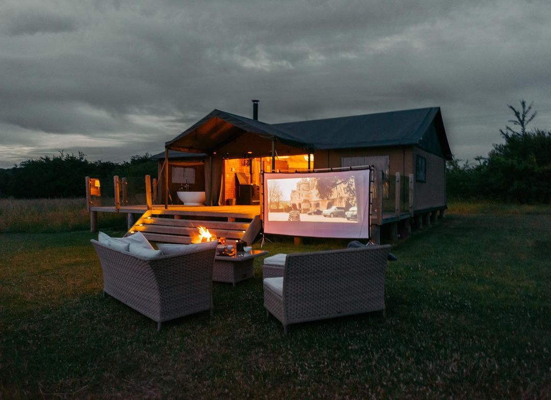 Embrace the magic of Nest Glamping with an eco-friendly outdoor movie setup: a screen, two wicker chairs, and a fire pit create a cozy retreat in front of the cabin as dusk settles in.