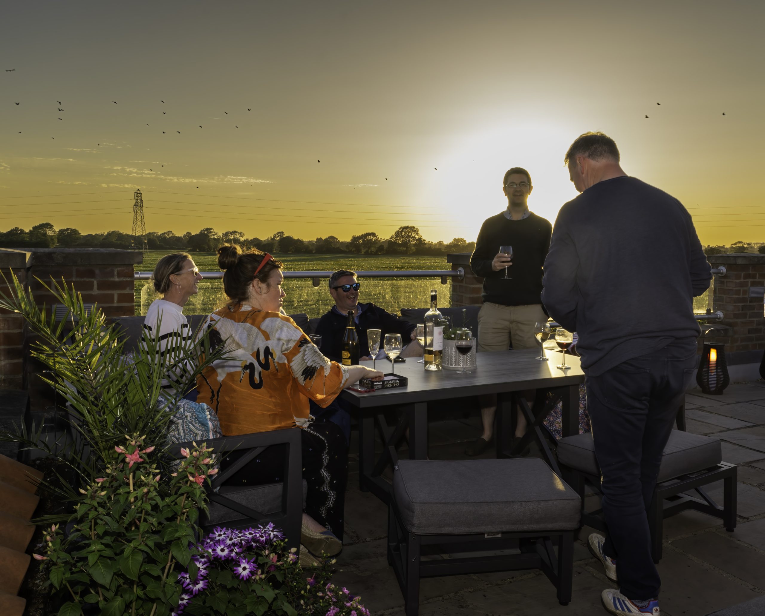 Five people are gathered around a table on a patio at sunset, enjoying drinks and conversation, as they share their New Year's resolutions.