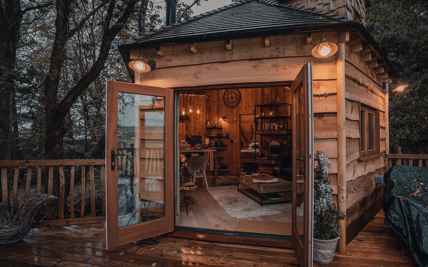 Cozy woodland cabin with open glass doors, warmly lit interior, nestled amidst trees on a wet wooden deck.