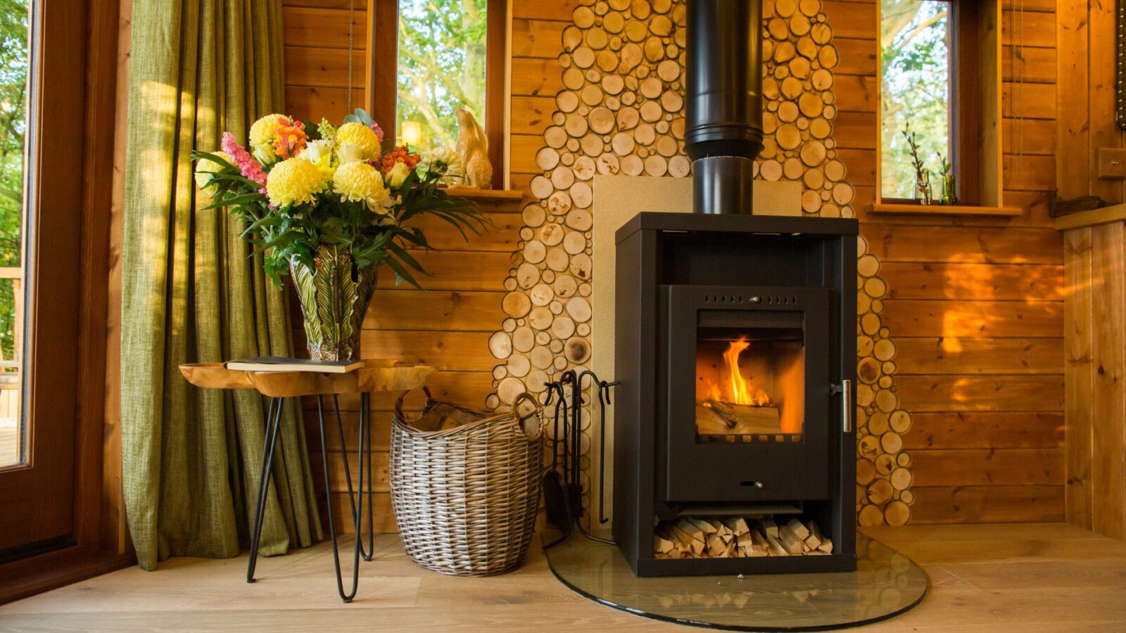 Nestled in the charm of Woodland Chase, this cozy wooden cabin interior features a lit wood-burning stove, along with a basket and a vase of flowers resting on a small table.