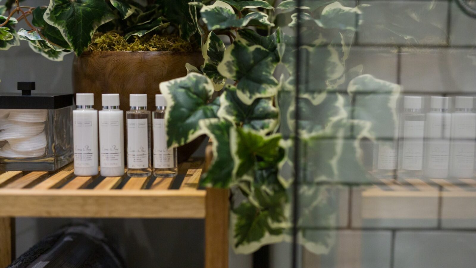 Small bottles on a wooden shelf in this cozy treehouse, surrounded by green plants, beside glass tiles and a stack of black towels.