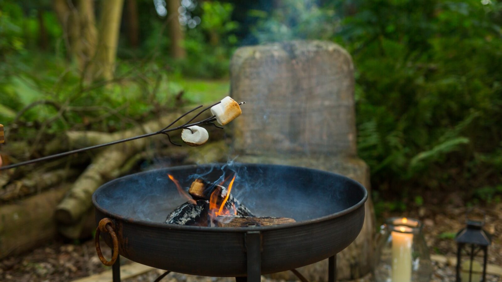 A fire pit with marshmallows on skewers being roasted, set in a serene woodland chase. Surrounded by lush greenery and the warm glow of a candle, a stone seat nestled nearby invites relaxation under the canopy, as if one might stumble upon a hidden treehouse just beyond.