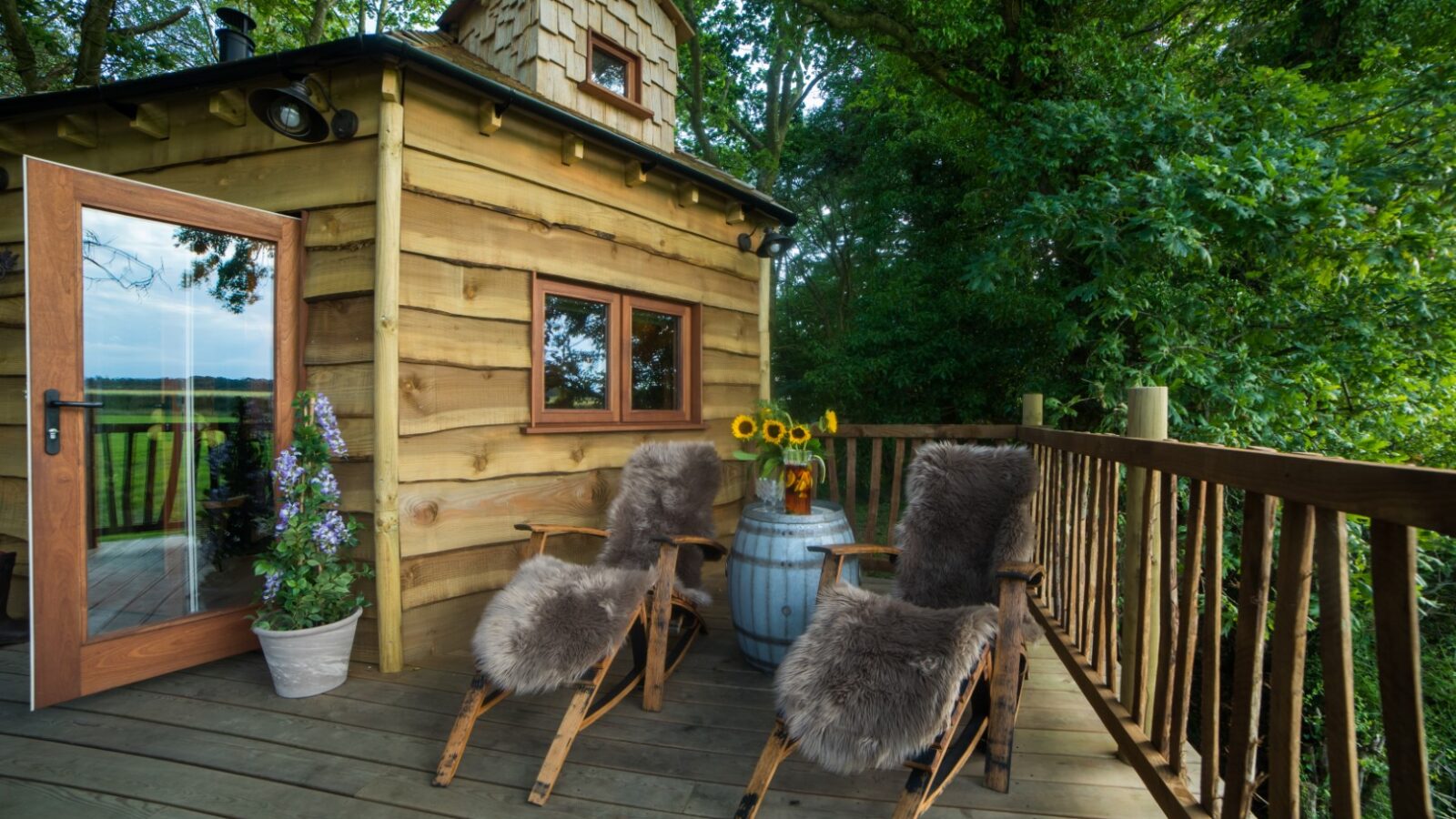 A cozy Treehouse retreat featuring a wooden deck with fur-covered chairs, a charming table adorned with potted flowers, and an enchanting lush forest view—a true Woodland Chase escape.