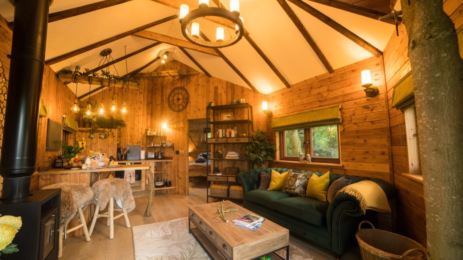 Rustic cabin interior with wooden walls, green sofa, coffee table, and kitchen area exudes a treehouse charm. Ceiling lights illuminate the cozy and warm atmosphere of this woodland chase haven.