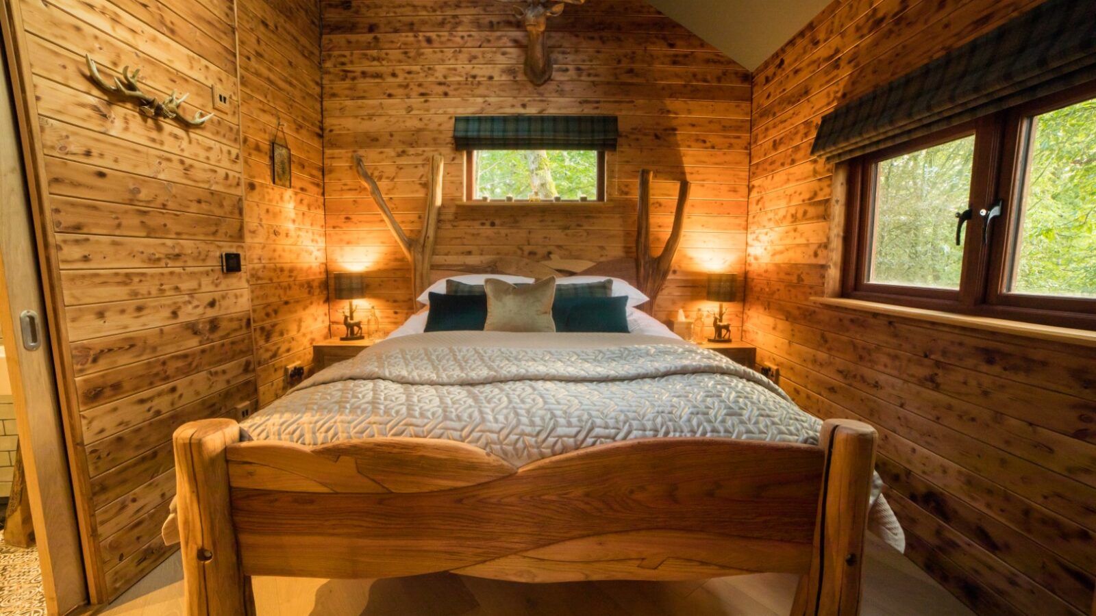 Cozy wooden cabin bedroom with a double bed, antler decor, and warm lighting. A large window on the right side offers a glimpse of the enchanting Woodland Chase outside.