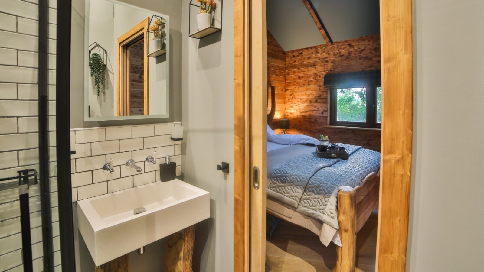 A rustic bathroom with a small sink and mirror evokes a woodland chase, seamlessly transitioning into a cozy wooden bedroom reminiscent of a treehouse, featuring a bed adorned with a quilted blanket.